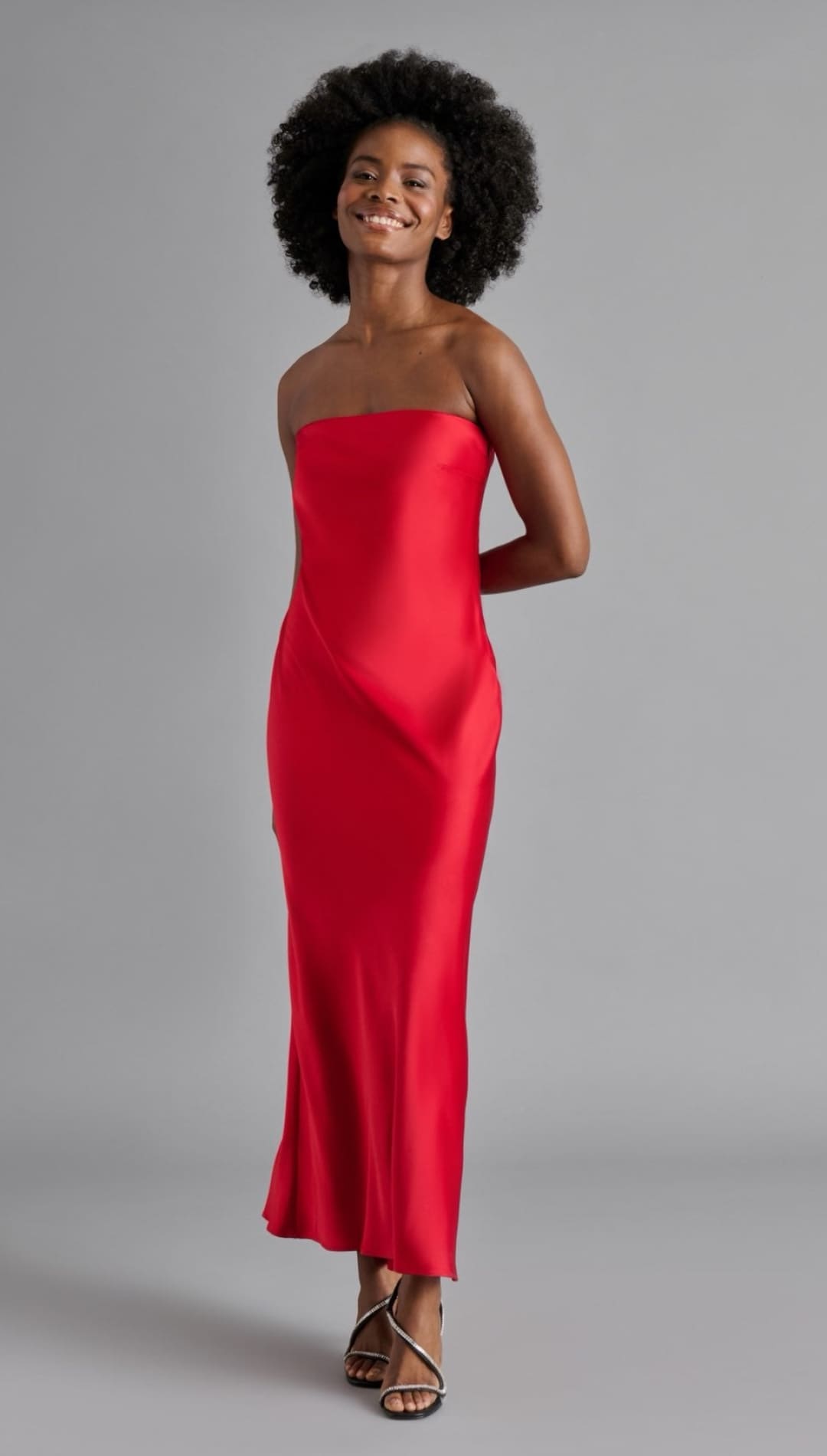 A smiling woman in a red strapless satin maxi dress and sparkly high-heeled sandals.