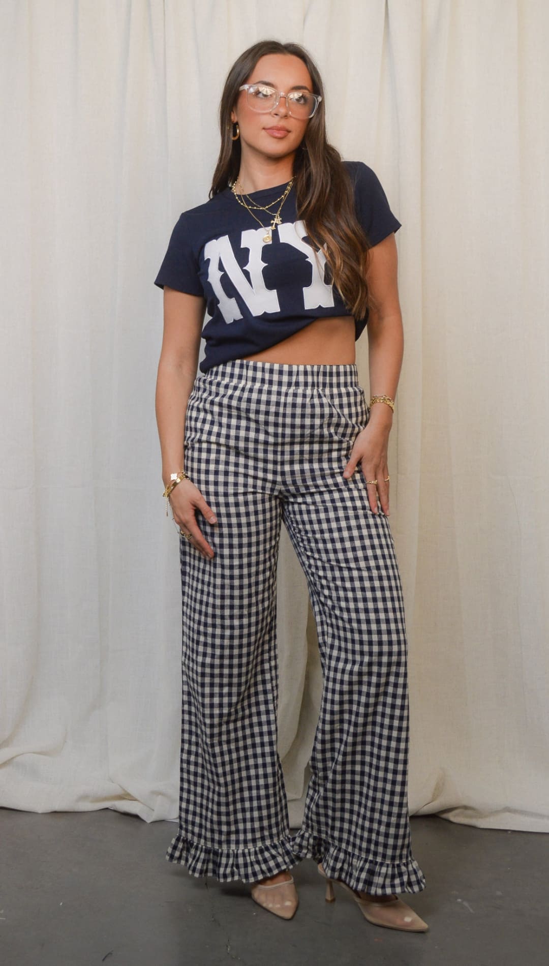 A woman modeling a casual outfit consisting of a cropped navy blue ’NY’ graphic t-shirt, black and white gingham wide-leg pants with ruffled hems, and nude pointed-toe heels.