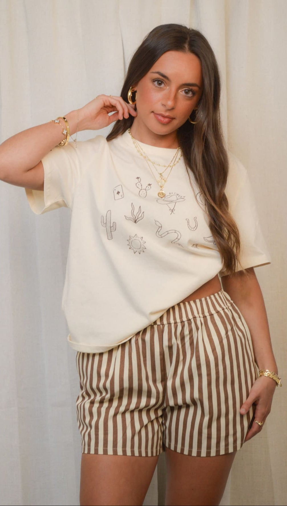 A woman models a cream-colored t-shirt with thin line drawings and brown and white striped shorts.