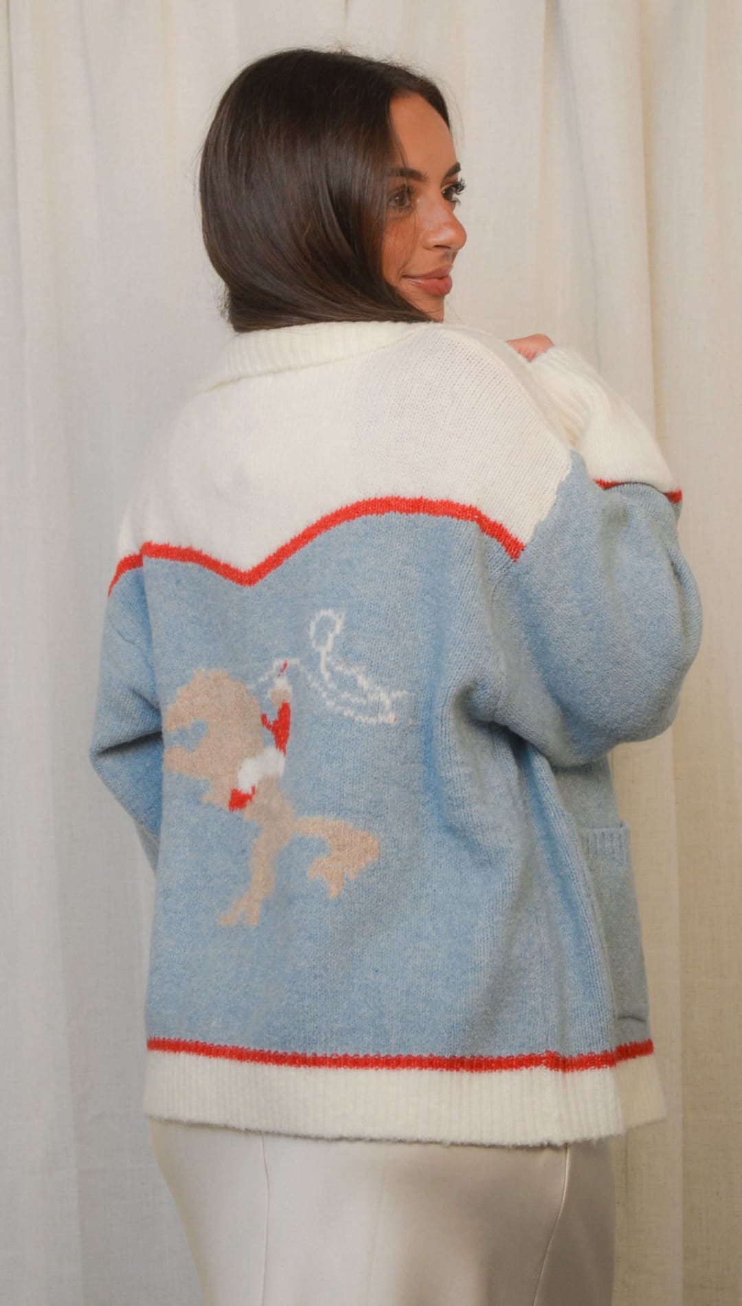 A woman wearing a light blue and white color-block cardigan with a red trim and a white deer-like figure on the back.