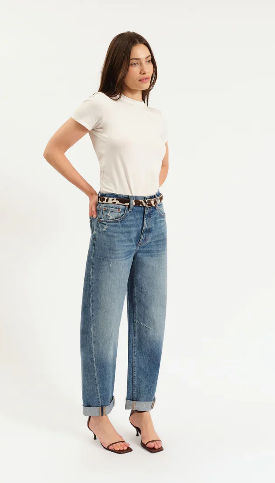 A woman in a cream t-shirt, blue jeans, a leopard print belt, and black kitten heels.