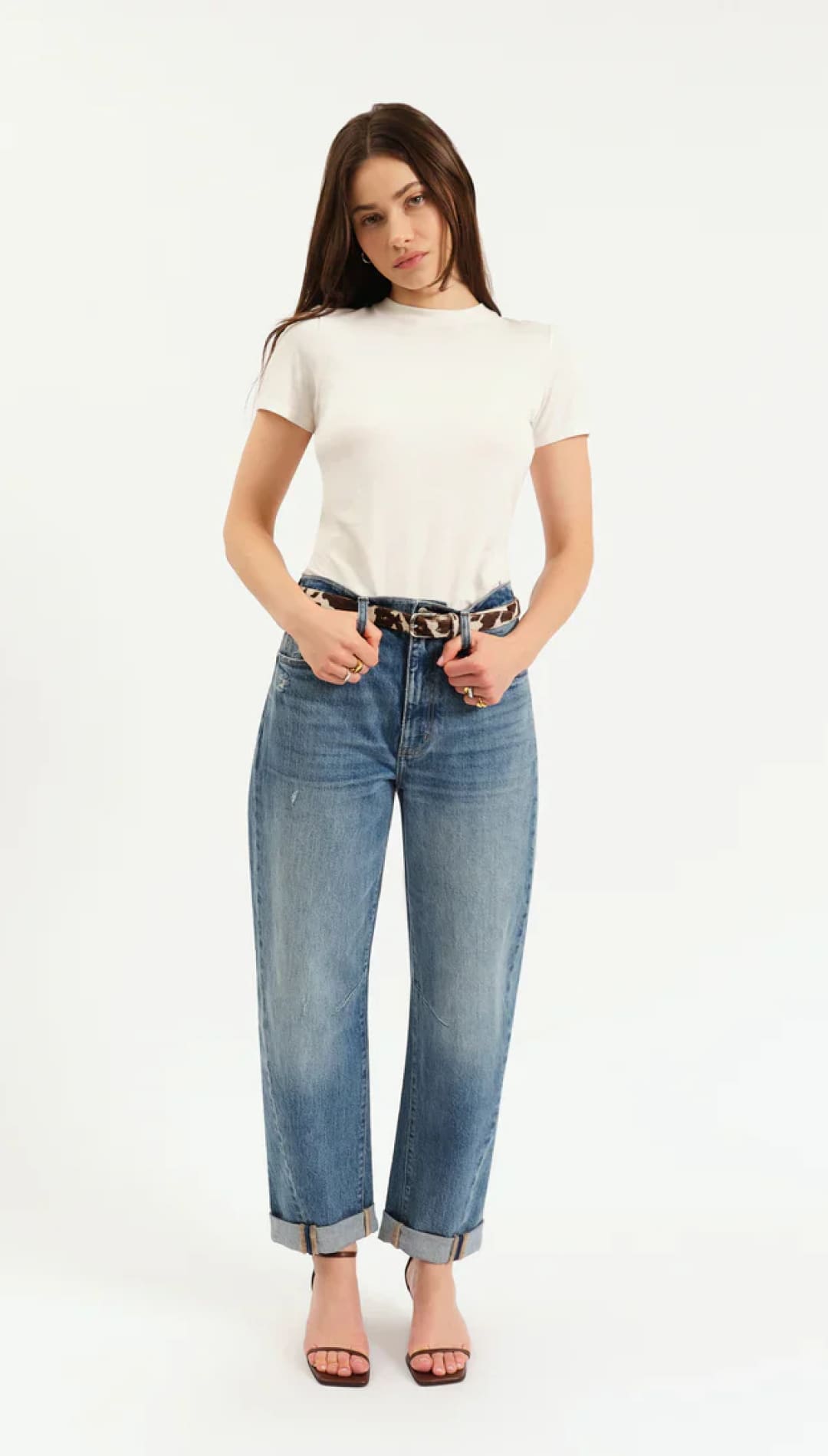 A woman wearing a white short-sleeve t-shirt, blue cuffed jeans, a leopard print belt, and brown heeled sandals.