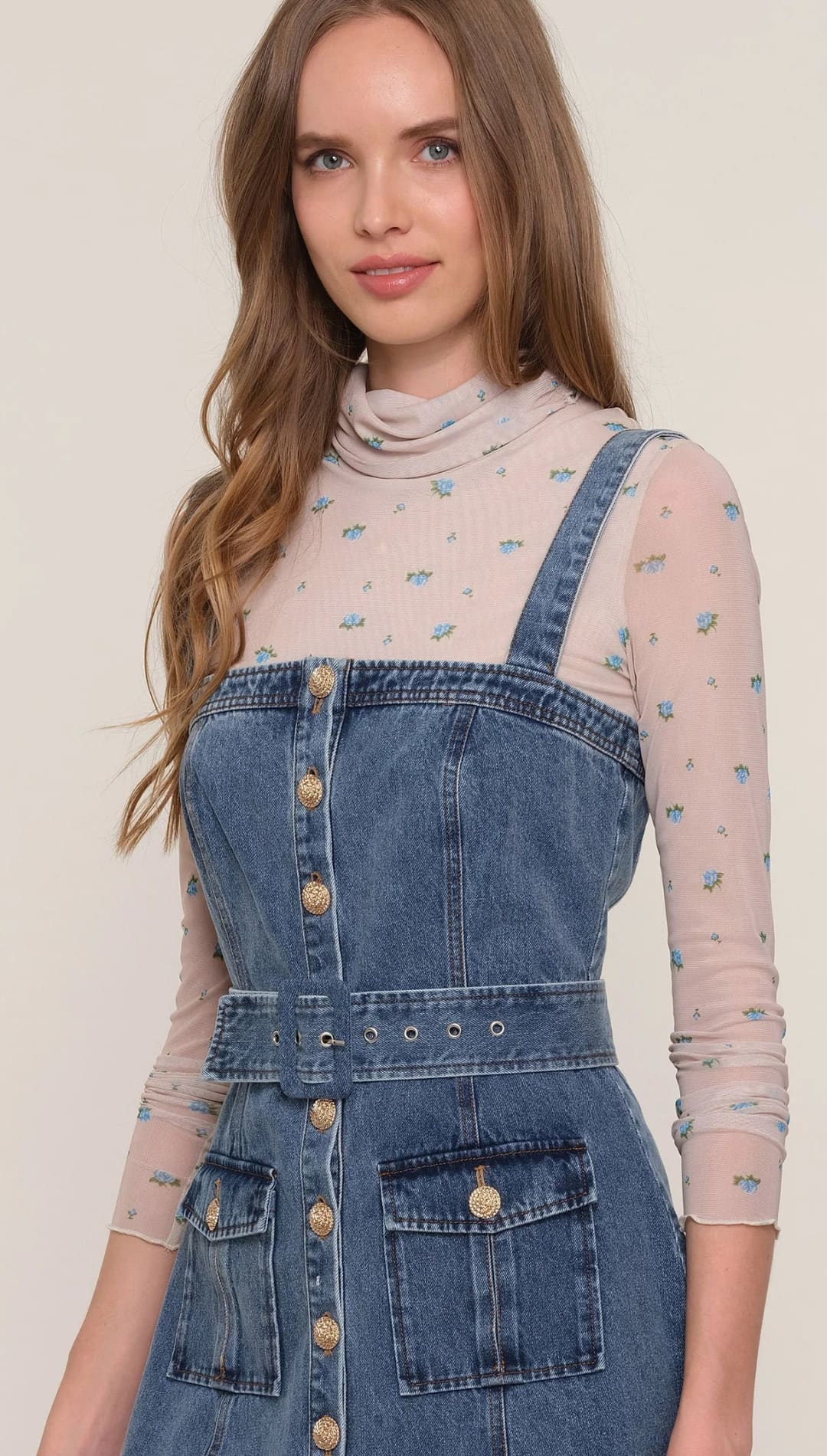 A woman wearing a denim overall dress over a sheer floral long-sleeve top.