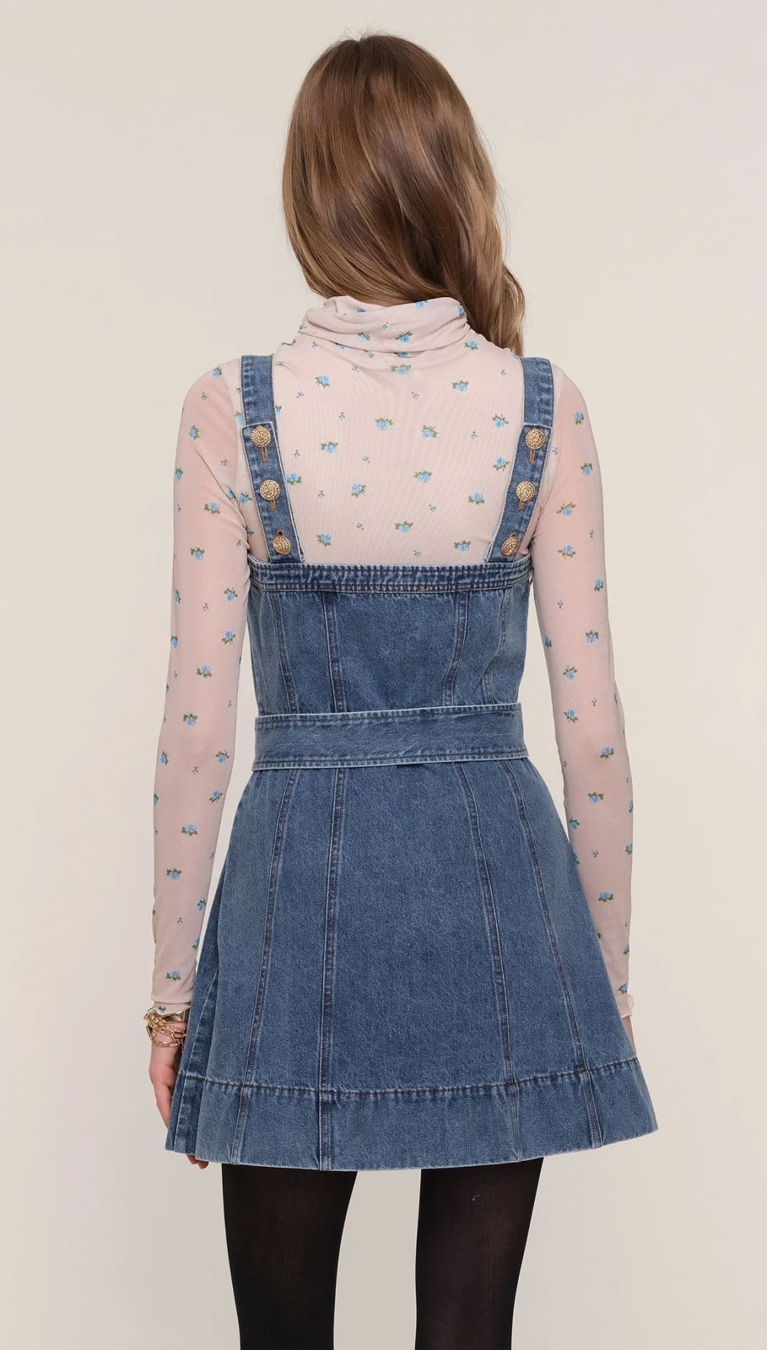 A woman wearing a denim overall dress over a sheer, long-sleeved turtleneck shirt.
