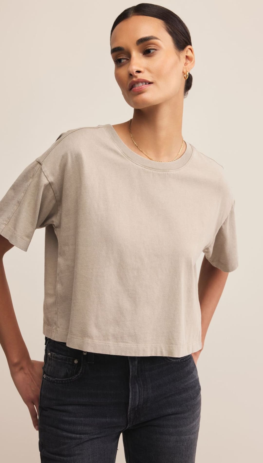 A woman modeling a loose-fitting, cropped light brown t-shirt with short sleeves and dark-wash jeans.