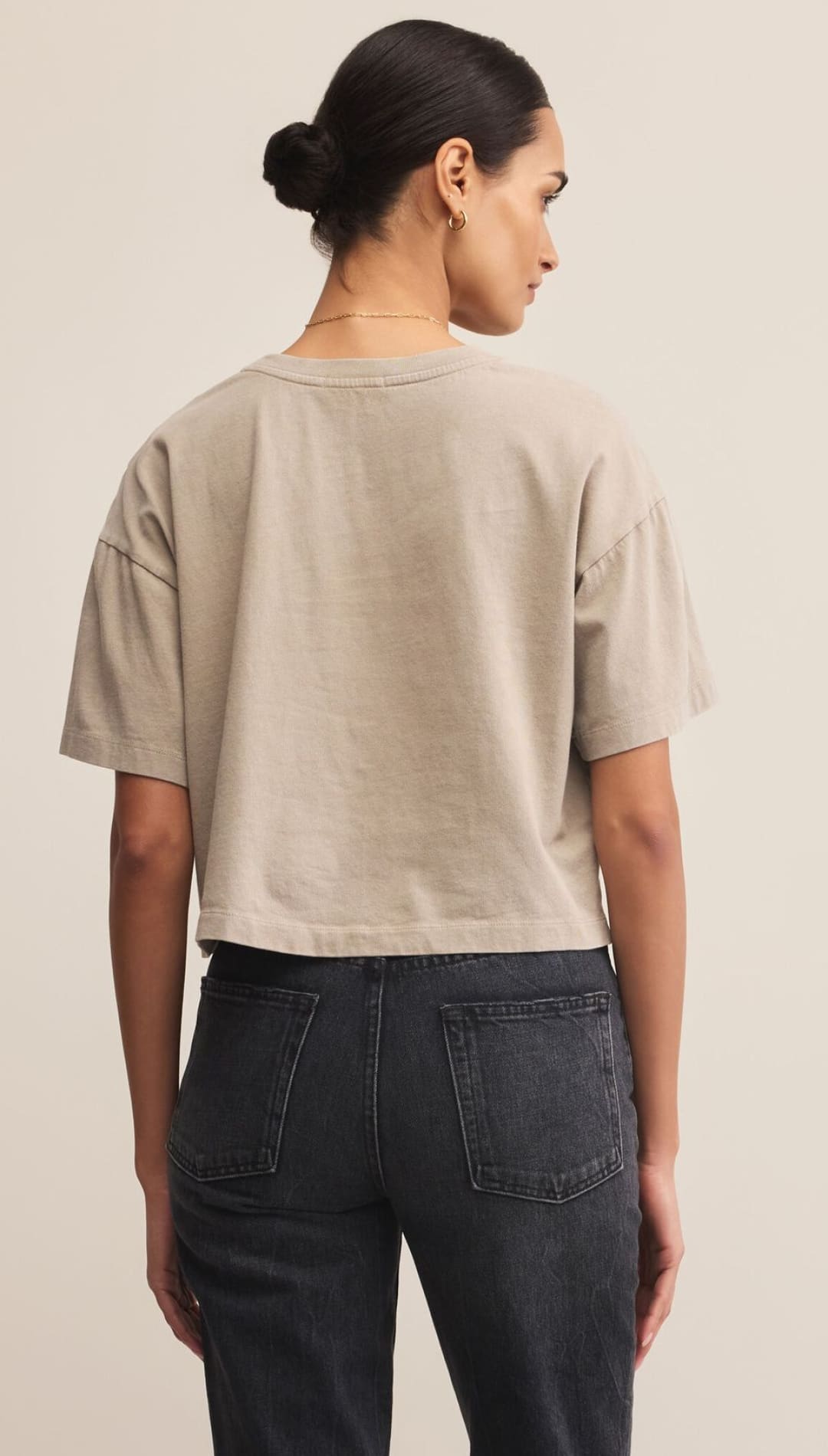 A woman from behind, wearing a loose-fitting, cropped tan t-shirt and dark-wash jeans.