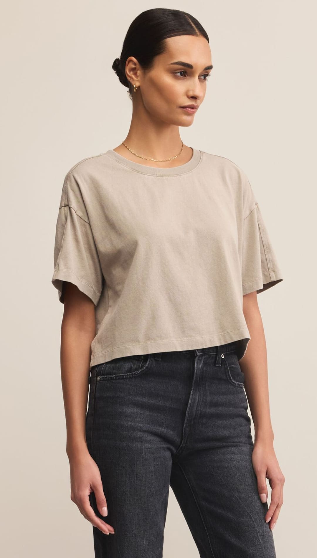 A woman models a relaxed-fit, light brown cropped t-shirt and dark grey jeans.