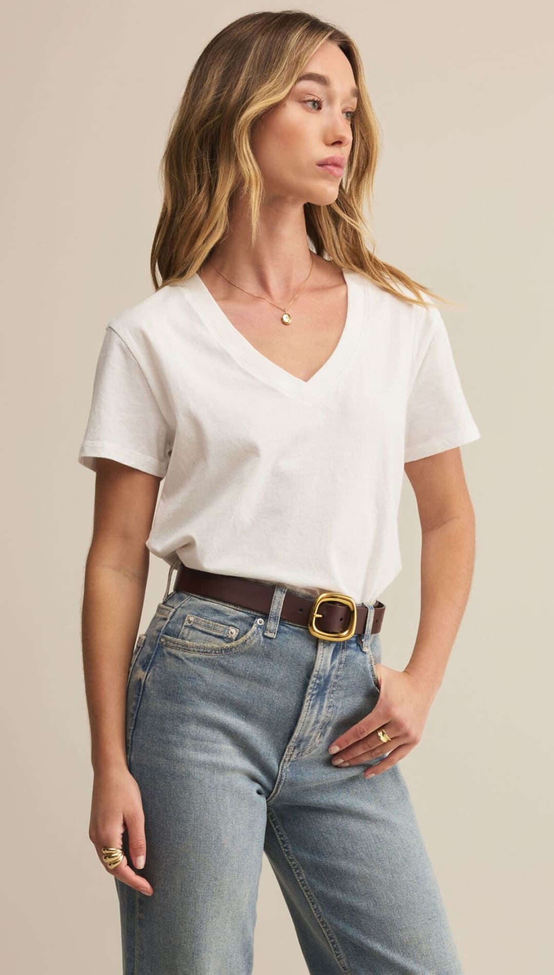 A woman wearing a white V-neck t-shirt, light-wash jeans, and a dark brown belt with a gold buckle.