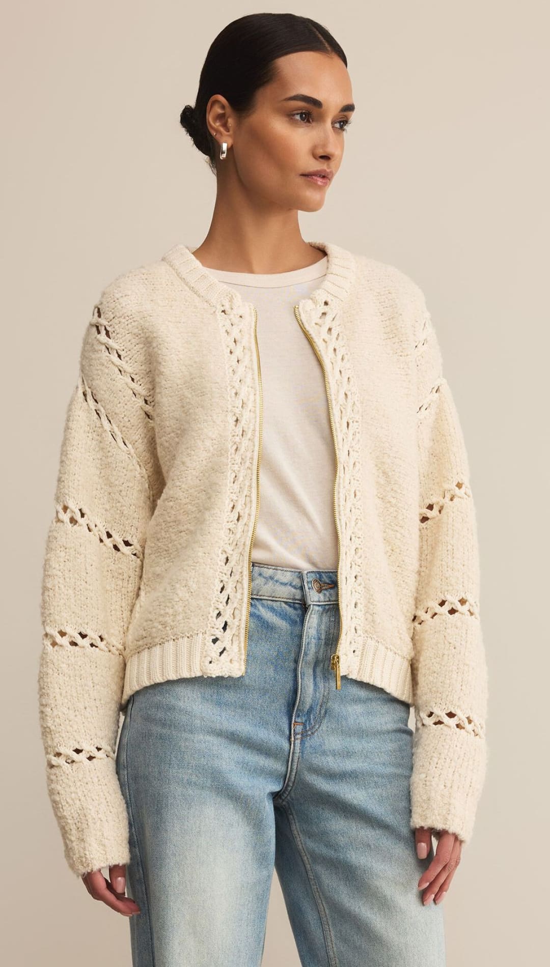 A woman wearing a cream-colored, open-front cardigan with textured knit and horizontal openwork details, paired with light-wash blue jeans.