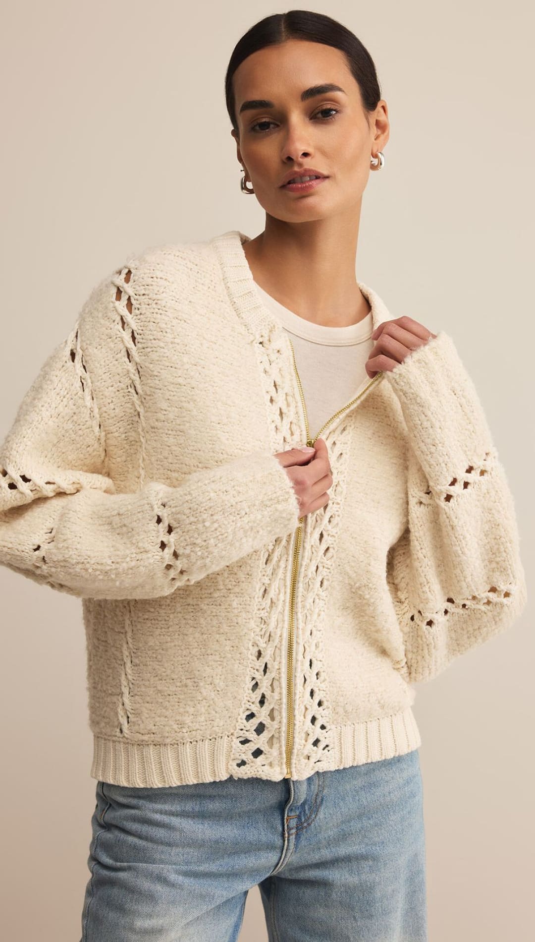Woman wearing a cream-colored, textured knit cardigan with openwork details and a zipper closure.