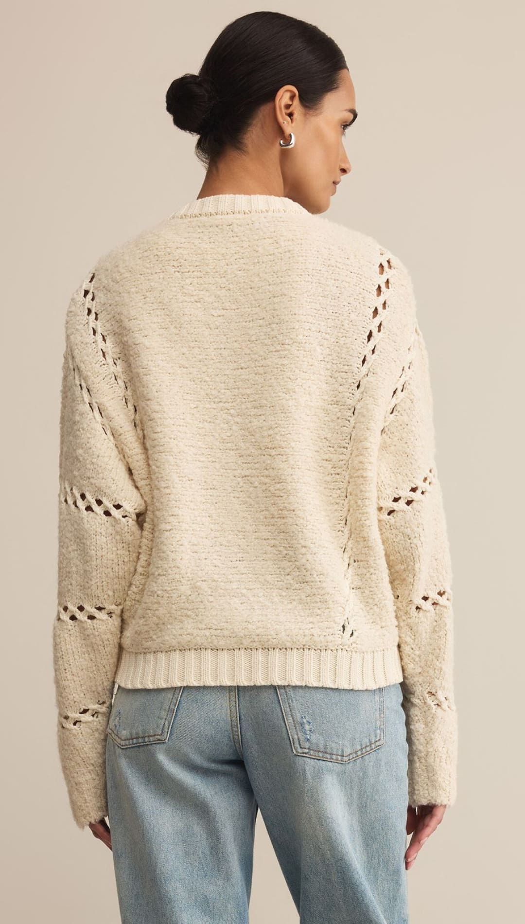 Women’s cream-colored long-sleeved knit sweater with open-knit details and a ribbed hem, paired with light wash denim jeans.