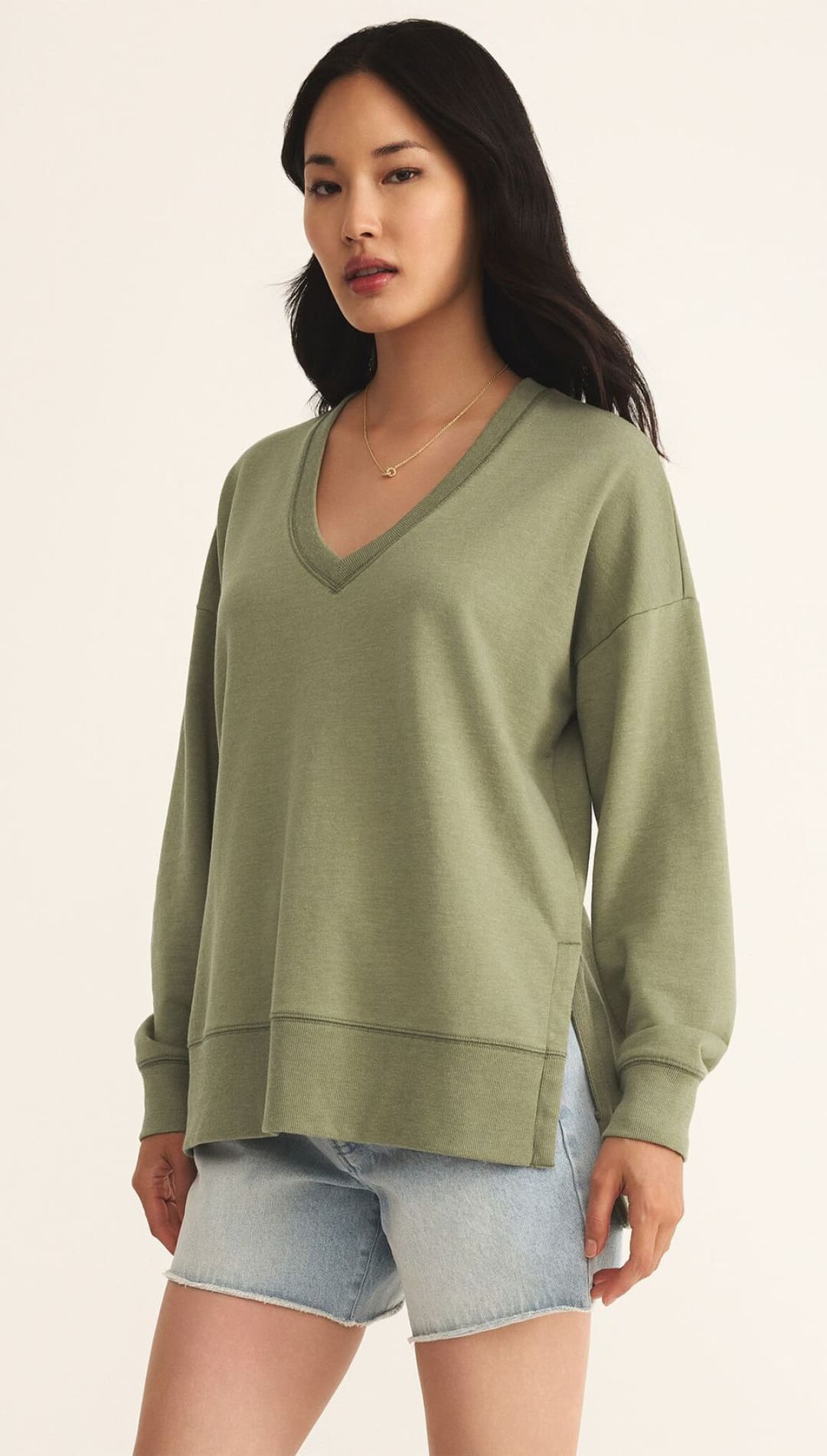 A woman wearing a relaxed-fit, long-sleeved, deep V-neck olive green sweatshirt with side slits and light-wash denim shorts.
