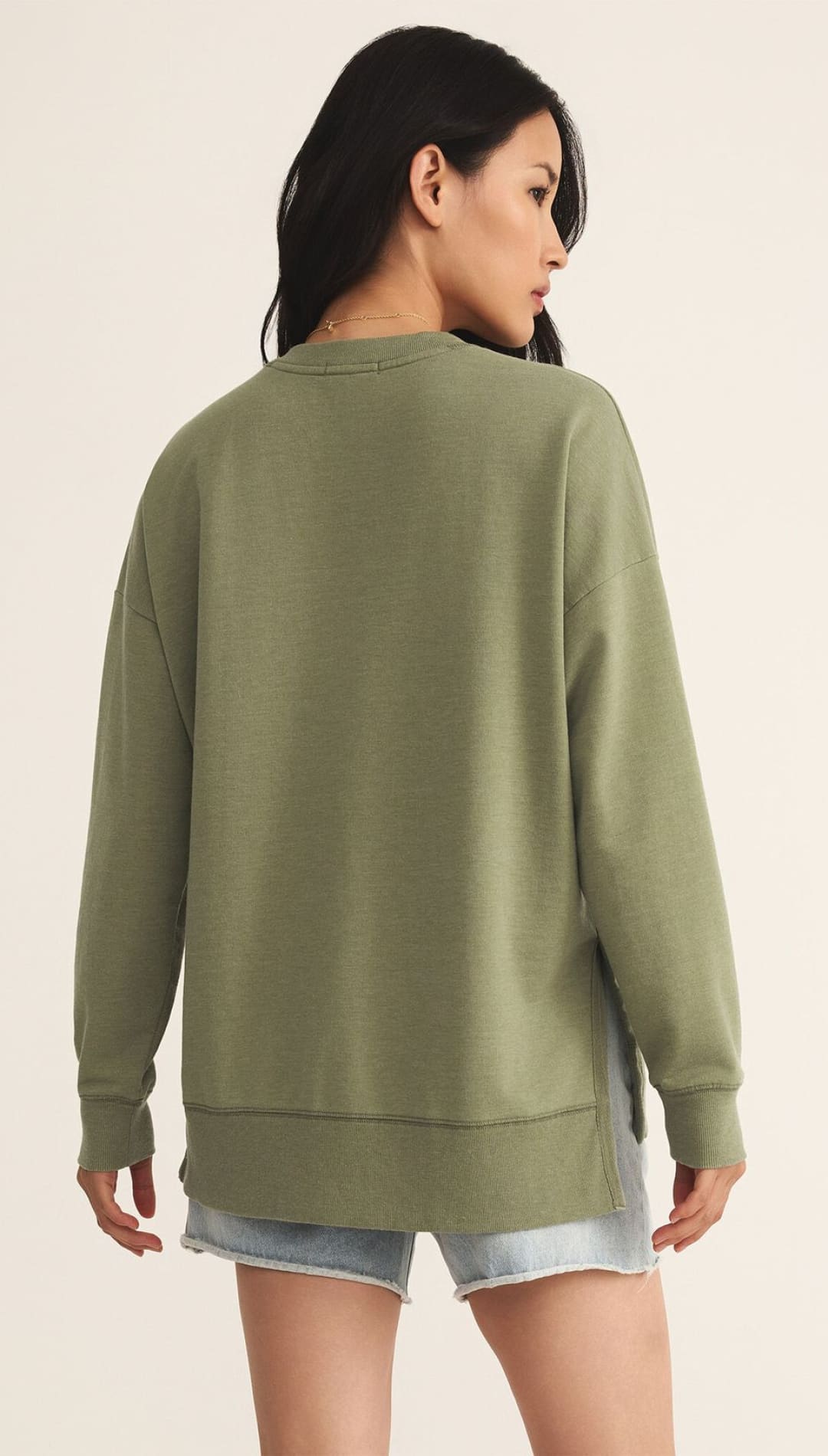 A woman wearing a relaxed-fit olive green long-sleeved sweatshirt with a ribbed hem and side slits, paired with light-wash denim shorts.