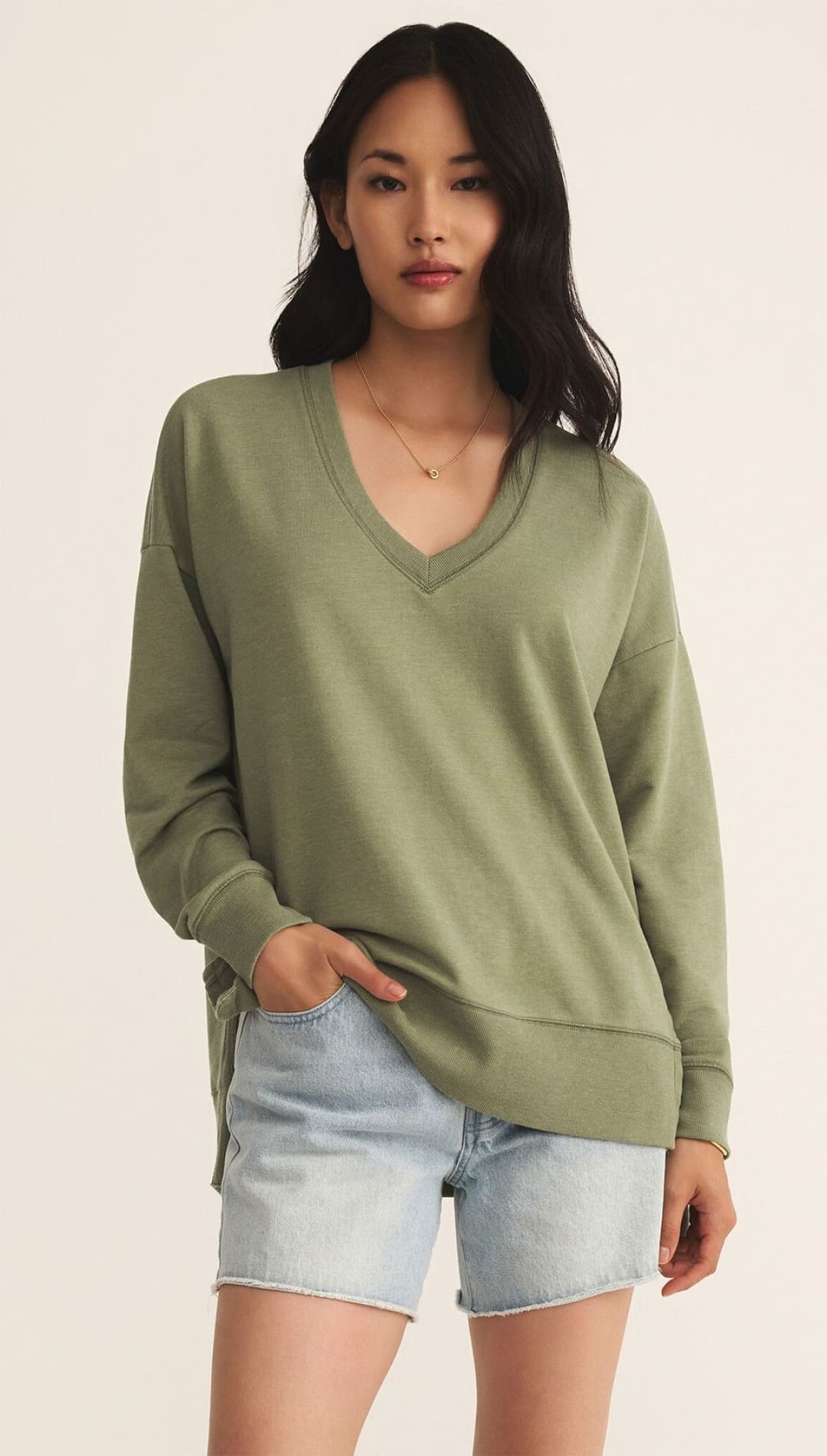 A woman wearing a relaxed-fit, olive green V-neck long-sleeve top and light-wash denim shorts.