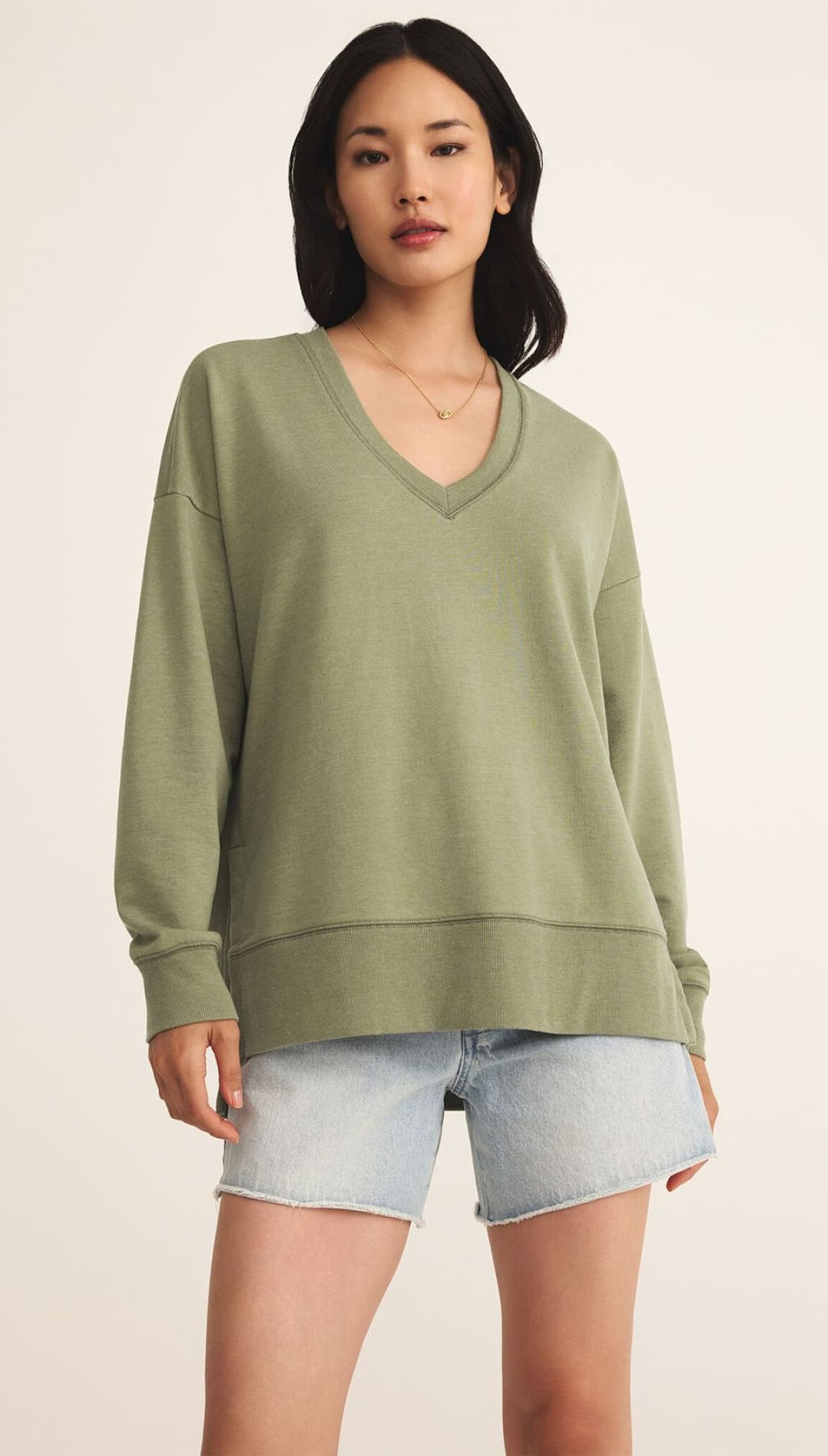 A women’s long-sleeved, loose-fitting, v-neck sweatshirt in a muted olive green, paired with light-wash denim shorts with frayed hems.